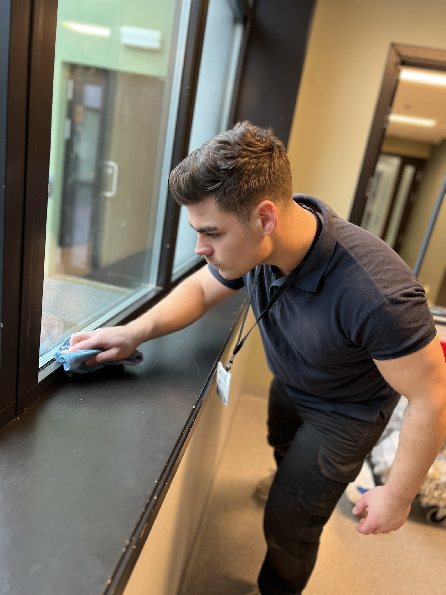 A person in a blue uniform and gloves cleaning a large window with a squeegee, reflecting clouds and sunlight.